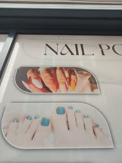 The image shows a close-up of a window display featuring a sign for a nail salon. The sign has the words "NAIL PO" visible in large, stylized font. Below the text, there are two oval-shaped graphics. The top graphic displays a close-up of hands with manicured nails, some painted orange and some with jewelry. The bottom graphic shows a close-up of feet with pedicured toes, painted in a bright blue color. The window appears to be made of a mesh material. The overall impression is a clean and professional advertisement for a nail salon. This advertisement is located on a window in the city of Rosenheim, Germany.