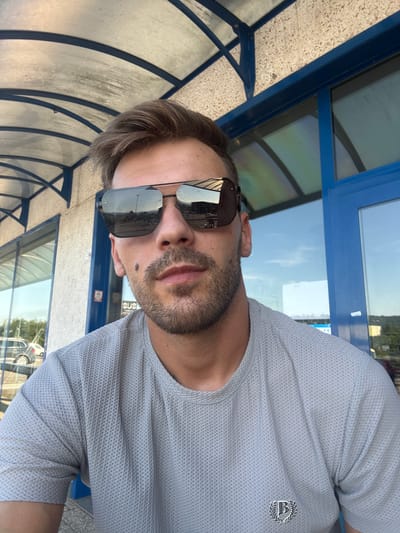 A man with brown hair and a beard, wearing sunglasses and a light grey t-shirt with a crest logo, is photographed from the chest up. He is outdoors, under a covered walkway with a blue frame and a textured ceiling. The walkway appears to be outside a building with large blue-framed windows. The background shows a sunny sky and some foliage. Reflections in the man's sunglasses show a landscape with a road and buildings. The location is identified as Castelletto sopra Ticino, Italy.