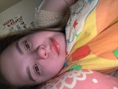 A person with brown eyes, fair skin, and glossy lips is partially covered by colorful bedding. A portion of a beige patterned garment is visible near the person's head. A poster is affixed to the wall behind them, featuring the words "Happy Birthday!" and some smaller images. The angle of the photo suggests the camera is slightly above the person.