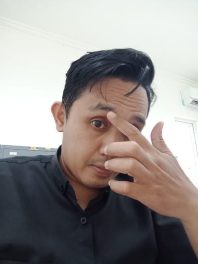 A man with dark hair styled upwards and wearing a dark button-down shirt poses indoors. He's touching his forehead with his index finger, and the rest of his hand obscures part of his face. His expression is serious. Background details include a white wall, part of a gray filing cabinet, and a window with an air conditioning unit. The lighting is bright and even.
