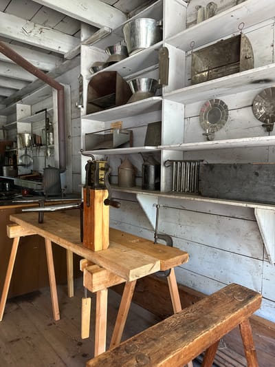 The image depicts the interior of a workshop or storage area, likely a historical exhibit, filled with antique tools and kitchenware. The room is constructed with rustic wooden planks for both walls and ceiling. Shelves line the walls, displaying a variety of metal containers, jugs, and implements, many of which appear to be made of tin or aluminum. Notable items include large buckets, a butter churn with a wooden base and metal mechanism, a candle mold, and several dark, rectangular boxes. A wooden workbench with a vise and sawhorses occupies the foreground, supporting the butter churn. A wooden stool is partially visible in the lower right corner. A cast-iron stove is positioned against the back wall, with a large metal pipe extending upwards. The overall atmosphere suggests a preserved or recreated historical setting, possibly related to domestic life or early industrial practices. The image does not contain any information that would allow for its specific location in New York City, United States of America.