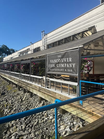 The image shows The Vancouver Fish Company, a restaurant and bar located in Granville Island, Vancouver, Canada. The restaurant features an outdoor seating area with a covered patio, decorated with hanging flower baskets. The building is situated on the waterfront, with a rocky shore visible below a wooden boardwalk. A clear blue sky is visible in the background.