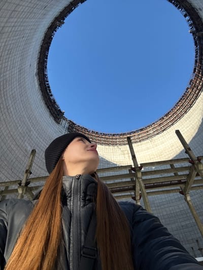 The image presents a low-angle perspective from inside a vast, cylindrical structure, looking upwards towards a clear blue sky. The original image is rotated 90 degrees counter-clockwise, making the sky appear on the left and the ground on the right from the viewer's immediate perspective. If viewed upright, the image shows a person standing inside what appears to be a large, abandoned cooling tower.

On the right side of the frame, partially obscured by the foreground structure, stands a person, likely female, observed from the chest up. She has long, straight, reddish-brown hair that cascades down her back, visible on the right side of the image. She is wearing a dark, possibly black, knitted beanie and a dark, puffy jacket with a zipper detail on the chest area. A dark shoulder bag strap is visible across her right shoulder. Her head is tilted slightly upwards and her gaze is directed towards the vast opening above, suggesting contemplation or observation of the immense scale of the structure and the sky. Her profile shows a defined jawline, lips slightly parted, and a focused expression.

The dominant feature of the scene is the interior of the massive concrete structure, which encircles a perfect, bright blue circle of sky. This structure's walls are made of light-colored, possibly off-white or light gray, concrete panels that appear to be textured or patterned in a grid-like fashion, consistent with the interior of a cooling tower. At the very top edge of the structure, where it meets the sky, exposed rebar or metal scaffolding is visible, indicating either an unfinished construction or significant deterioration over time.

To the right and slightly behind the person, there are additional industrial-looking support beams or scaffolding. These are constructed from lighter-colored concrete and dark metal bars, forming a complex lattice work that extends vertically, adding to the sense of an industrial or derelict setting.

The sky is uniformly bright blue and completely cloudless, suggesting a clear day. The lighting is strong, casting subtle shadows and highlighting the textures of the concrete and the person's clothing. No discernible text is visible anywhere in the image. The overall impression is one of immense scale and quiet observation within an imposing, man-made environment.