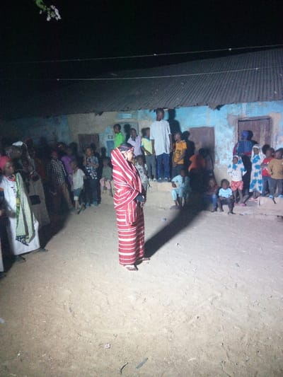 A woman in a red and white striped robe stands in a dirt yard surrounded by a crowd. The crowd includes men, women, and children. The woman is facing right. Behind the crowd is a building with a corrugated metal roof. The scene appears to be at night or in dim light. Some people are standing on the steps or platform of the building, while others are on the ground. The location may be a village or rural area.