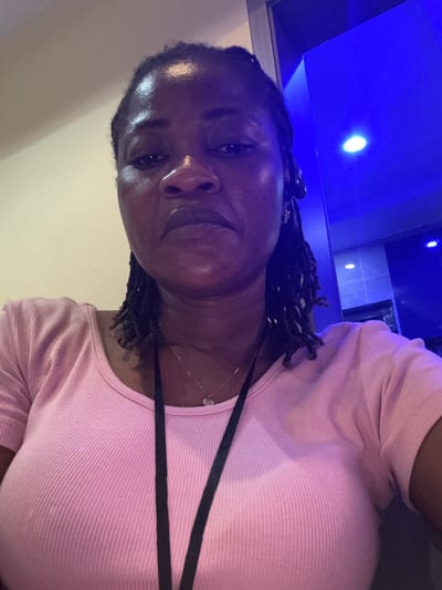 The media file shows a close-up, sideways shot of a Black person wearing a pink ribbed top. The individual has dark, braided hair and is wearing a thin silver necklace with a small pendant. They are looking towards the camera with a relaxed expression. The background is dimly lit, with a prominent blue light source visible in the upper left portion of the image, suggesting an indoor setting with artificial lighting, possibly at night. The overall lighting casts some shadows and highlights on the person's face and clothing. There are no visible objects or text in the frame. The context provided places the scene in Ajara, Nigeria.