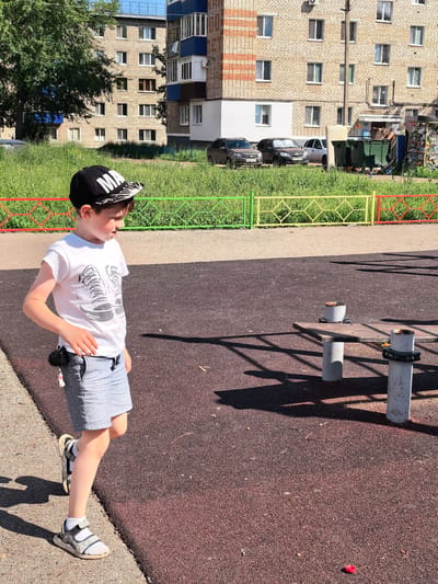 A young boy with light skin and brown hair is walking on a dark red playground surface. He is wearing a black baseball cap with white lettering that reads "MA," a white t-shirt with a graphic of two sneakers, and light blue denim shorts. He has on white socks and gray and white sandals. A dark shadow of a fence is cast across the playground to his right. In the background, there is a green grassy area and a multi-story brick building with numerous windows. To the left of the building, there is a large tree. In front of the building, there are several cars and large green trash bins. A colorful fence with green, yellow, and red sections separates the grassy area from the playground. A small red object is on the ground in the lower right corner of the image. The scene appears to be a residential area in the city of Sterlitamak, Russia, likely a courtyard with a playground.