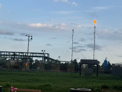 The image depicts an industrial site in Igwuruta, Nigeria, at dusk or dawn, under a cloudy sky. Prominent features include two tall flare stacks, one of which is actively burning off gas, emitting a bright orange flame. In the foreground is a grassy field, bordered by a chain-link fence, behind which sits various industrial equipment including interconnected metal structures and a blue tank. There is also a small shelter-like structure. Lampposts are scattered throughout the scene. The overall impression is of an active oil or gas processing facility.