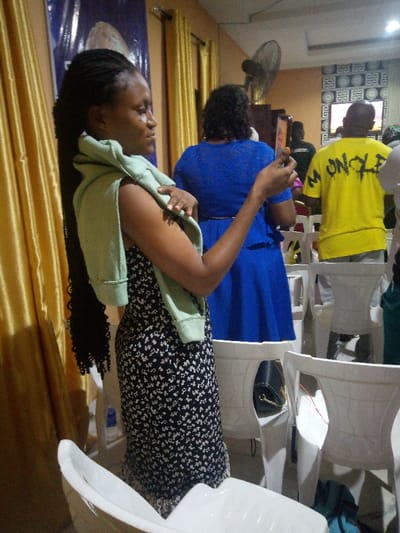 Inside a church-like venue, a woman in a floral dress and a light green sweater draped over her shoulders is using her phone, possibly taking a photo. Behind her, a woman wears a blue dress. Further back, a person in a yellow shirt with "M UNCLE" printed on it stands among others, seemingly watching a screen. White plastic chairs fill the space, and gold curtains hang on the left. A fan is visible on the back wall.
