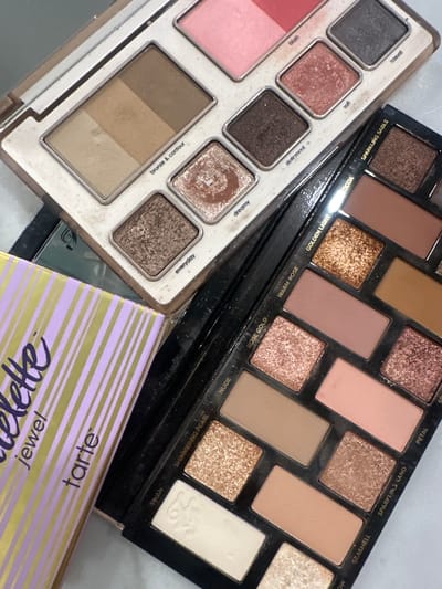 The image displays a close-up, top-down view of three distinct cosmetic palettes arranged on a light-colored, possibly white or off-white, surface. The location in the city of Monterrey, Mexico, cannot be determined from the visual evidence.
In the upper-left area, a rectangular cosmetic palette with a light beige frame is visible. This palette contains a long rectangular pan divided into three shades of matte brown for "bronze & contour", two square pans of blush in pink and coral tones, and five square eyeshadow pans. The eyeshadows include a shimmery light brown labeled "everyday", a shimmery copper labeled "dreamy", a dark matte brown labeled "dutchess", a shimmery peach labeled "soft", and a dark gray labeled "casual". The palette shows residue from use, with powder visible around the edges of the pans.
Below and to the right, a second cosmetic palette with a black casing is partially visible, oriented diagonally. This palette contains twelve pans of various eyeshadows and possibly a highlight. Visible shades include a shimmery gold labeled "Golden Light", a matte brown labeled "Cocoa", a shimmery rose gold labeled "Rose Gold", a matte nude labeled "Nude", a shimmery beige labeled "Shimmering Pearl", and a cream-colored pan labeled "Swan" which features an embossed swan emblem. Other visible shade labels are "Warm Rose", "Sparkling Sable", "Petal", "Sparkling Sand", and "Seashell". This palette also exhibits slight cosmetic powder residue.
On the far left, partially obscured by the other palettes, is a section of a lavender-colored box or container with vertical gold stripes. Text printed on this item clearly reads "tartelette jewel tarte", identifying it as packaging for a Tarte Cosmetics product.