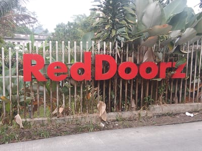 The image shows a bright red "Red Doorz" sign mounted on a metal fence. The sign is large and prominently displayed. Behind the fence, there are large tropical leaves, likely from banana or heliconia plants, and other lush green foliage. In the background, a building with white walls and a dark roof can be seen, suggesting a commercial or residential property. The fence itself is made of vertical metal bars with decorative scrollwork. The ground in front of the fence is covered with dirt, grass, and some fallen leaves, indicating an outdoor setting. The overall scene appears to be in a warm, possibly humid, climate. The specific location is identified as Megamendung, Indonesia.