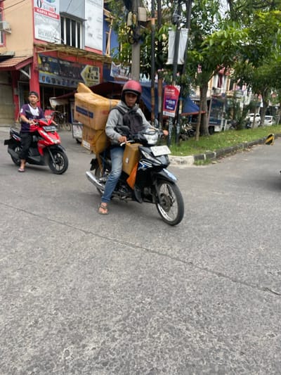The scene is a street in Margaasih, Indonesia, with a motorcycle loaded with cardboard boxes being driven by a man wearing a helmet. Another man is riding a red motorcycle in the background. The street is paved and surrounded by buildings and trees. The time of day is likely midday, as the shadows are short. The motorcycle is driving on the left side of the road.