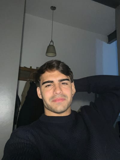 The image features a young man in a close-up shot, likely a selfie, looking directly at the camera with a relaxed expression and a subtle smirk. He has dark hair, prominent dark eyebrows, and brown eyes. Light stubble is visible on his chin and upper lip, and his lips are slightly parted. He is wearing a dark, ribbed knit sweater, possibly black or navy blue.
The setting is an indoor room with light-colored walls and a ceiling. In the upper left background, a hanging pendant light fixture with a bell-shaped, light-colored shade is visible. Further back, behind the man's head, some dark wooden furniture, possibly a shelf or cabinet, can be seen in the dimly lit environment. The subdued lighting suggests the photograph was taken in the evening or at night. The overall mood is casual and personal, likely taken in a residence in Avellaneda, Argentina. There is no visible text in the image.