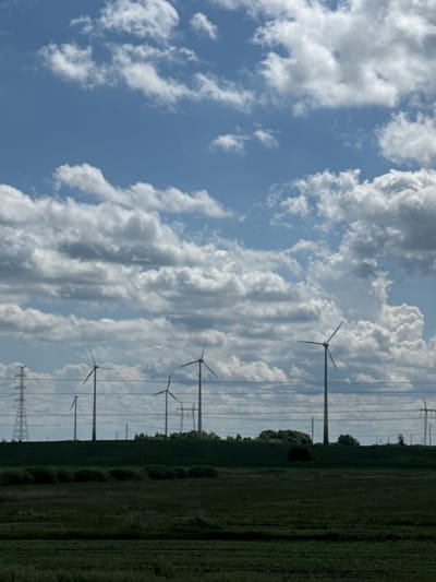 The image presents a rural landscape in Āres, Latvia, featuring renewable energy infrastructure and agricultural fields under a partly cloudy sky. The upper two-thirds of the frame display a bright blue sky with numerous white and grey cumulus clouds, varying in size and density, with some clear blue patches visible.
The mid-ground is characterized by a horizontal line of industrial elements. At least five large, three-bladed wind turbines are visible, silhouetted against the sky or partially illuminated. One prominent turbine stands on the far right, another near the center, and several others are positioned to the left and in the deeper background. A large lattice-steel electricity transmission tower is present on the far left, and multiple dark horizontal power lines stretch across the frame above the landscape elements. Additional, less distinct transmission towers are visible further in the background to the right.
The lower third of the image consists of dark green agricultural fields in the foreground, suggesting cultivation. These fields are separated from the background by a dark, elevated landmass, possibly a hill or embankment, that extends horizontally across the scene. Along the base or on the slope of this elevated land, a series of dark green, rounded bushes are distinctly visible in a linear arrangement.
No people, animals, or vehicles are present. The scene is illuminated by natural daylight, with diffused lighting conditions indicated by the cloud cover and muted tones of the landscape.