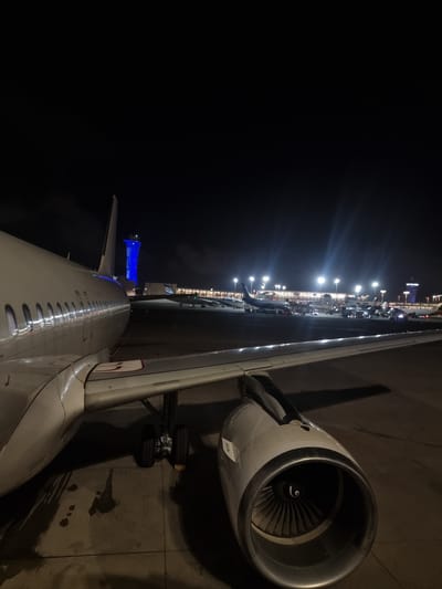 The image shows a nighttime view from an airplane window at an airport in Yagel, Israel. The aircraft's wing and engine are prominently featured, positioned on a tarmac with visible landing gear. Beyond the plane, a brightly lit terminal building extends along the horizon, interspersed with illuminated streetlights. A blue-lit structure is visible near the tail of the plane. The sky is a deep black.