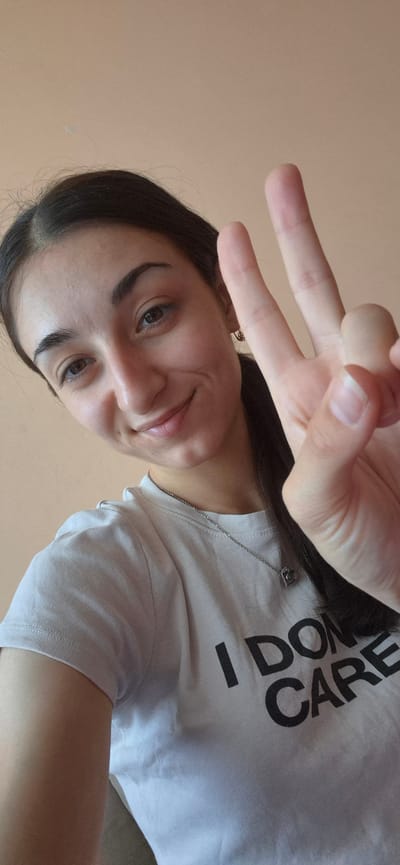 A young woman with long dark hair, wearing a white t-shirt with black text, takes a selfie. She is looking at the camera with a slight smile and her left hand is raised in a peace sign. The t-shirt has the words "I DON'T CARE" printed on it in large black letters. She is wearing a thin silver chain with a small pendant. The background is a plain, light brown wall. The lighting suggests it could be daytime, possibly indoors. There are no specific location cues other than the provided context of Silistra, Bulgaria. The overall mood is casual and lighthearted.