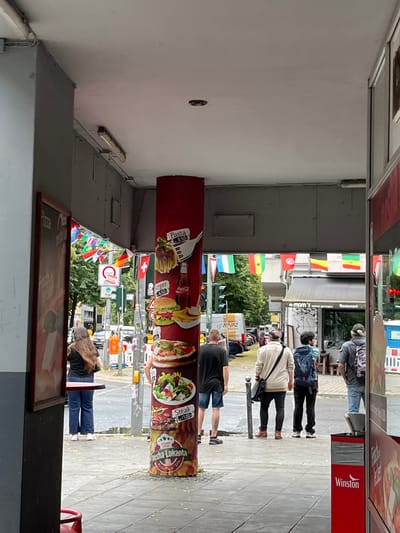 The image depicts a street scene in Berlin, Germany, with several people standing or walking along a paved sidewalk. A large, vertical banner advertising food, featuring images of pasta, burgers, pizza, and steak, hangs prominently in the foreground. The banner also displays prices like "Pasta 8,50€" and "Steak 13,00€". Small flags from various countries are strung across the scene, suggesting an international atmosphere. Traffic lights and street signs are visible in the background, indicating an urban environment. People are dressed casually, and the overall mood appears to be neutral, likely during daylight hours given the lighting. Some individuals are looking at their phones or towards the banner. A red and white sign with the word "Winston" is visible on the right side of the image. The presence of a restaurant or food establishment is implied by the advertising banner.