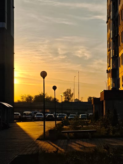 This image captures a sunset over a residential courtyard in Brest, Belarus. The scene is dominated by the warm, golden hues of the setting sun, which casts long shadows across the paved courtyard and illuminates the facades of two apartment buildings. On the left, a dark, modern building stands in silhouette, its windows reflecting the sky. On the right, a building with a textured facade glows in the sunlight, its many windows creating a grid-like pattern.
In the foreground, several cars are parked. A street lamp with a spherical head stands prominently, its dark pole contrasting with the bright sky. Another, smaller street lamp is visible further back. The paving stones of the courtyard are laid in a patterned design, with some areas brightly lit by the sun and others in deep shadow. A simple wooden bench sits in a patch of overgrown grass and bushes, bathed in the golden light. In the background, a bridge or elevated walkway crosses the scene, with trees and smaller buildings visible beyond it.
The sky is a gradient of soft oranges and yellows, with wispy clouds adding texture. The overall mood is peaceful and serene, with the setting sun creating a sense of warmth and tranquility. The time of day is clearly late afternoon or early evening, as indicated by the low angle of the sun and the long shadows. There are no people visible in the image. There is no visible text in the image.