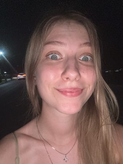 This is a close-up, low-light, selfie-style photo of a young woman with blonde hair and blue eyes. She is looking directly at the camera, with a slight smile. Her face is illuminated by artificial light, while the background is dark and out of focus, with some distant lights suggesting a nighttime scene. She is wearing a necklace with a small airplane pendant. The overall setting appears to be outdoors, given the darkness and distant lights. The image is oriented sideways.