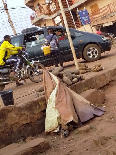 A bustling street scene shows a man in a yellow jacket on a motorbike, next to a dark green car. Another man in a striped shirt holds a yellow basket, seemingly interacting with the car's occupants. A pile of yams sits on the sidewalk, partially covered by a brown and cream tarp where two pigeons nestle underneath. A blue "Dangote Cement" sign hangs in the background, alongside multi-story buildings with balconies and intricate designs. The setting depicts a busy marketplace or roadside vendor area.