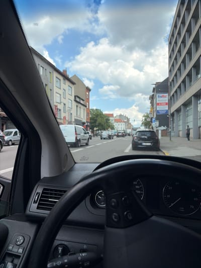 The image shows a view from the driver's seat of a vehicle, looking forward and slightly to the left. The scene is a street in Schaerbeek, Belgium. The weather appears to be cloudy, with patches of blue sky visible.
Prominently featured are the car's dashboard and steering wheel on the right side of the frame. The dashboard includes a tachometer and a speedometer, indicating the car is likely in motion. The steering wheel has controls, and part of the car's interior, including an air vent, is visible.
Outside the car, a street lined with multi-story buildings can be seen. Some buildings have balconies, and windows are visible. A few cars and vans are present on the street, including a white van with text on its side and a black car with a license plate. A sign for "BigMat" is also visible on one of the buildings. Trees are scattered along the street. The image seems to be taken during the daytime.
The perspective suggests the car is driving or stopped in traffic on a city street. There are no people clearly identifiable as subjects of the image, but a distant figure can be seen standing on a sidewalk. The overall impression is a snapshot of urban street life in Belgium.