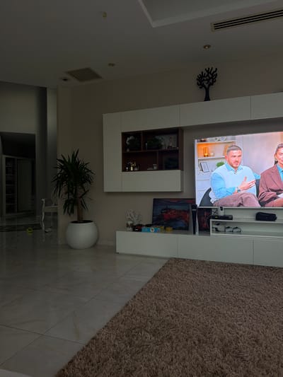 This media file depicts an indoor living space, likely in Kyrenia, Northern Cyprus. The scene is dominated by a large, modern entertainment unit. Mounted on the wall is a large flat-screen television displaying an image of two individuals, a man and a woman, seated. The man, in the foreground, is wearing a light blue shirt and appears to be speaking. The woman is dressed in a brown top. Below the television, the entertainment unit features several shelves and compartments. One open shelf displays decorative items and what appear to be framed photographs. Another section has closed cabinetry. To the left of the television, a black, decorative tree-like sculpture is mounted on the wall.

Further into the room, a large, shaggy, brown rug covers a section of the tiled floor. A tall green plant in a white, spherical pot stands near the rug. The overall lighting suggests it is daytime, but the interior setting creates a cozy ambiance. Other details include a white console table with some decorative items and a glimpse of a kitchen area or another room with appliances in the background. The image is captured from a slightly elevated, rotated perspective, indicating the photographer was standing or positioned to capture the scene from an unusual angle. There is no visible text in the media file.