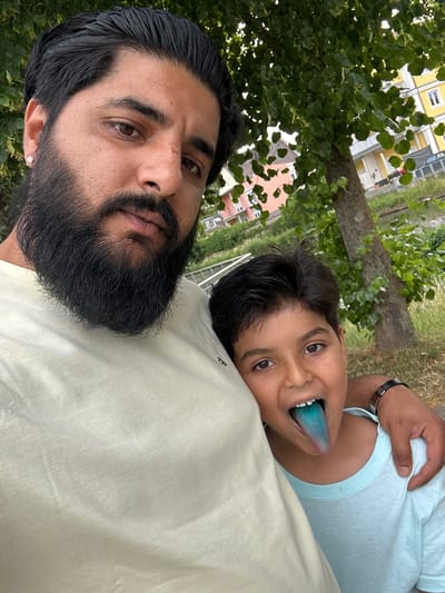 Two individuals, an adult male and a child, are captured in a selfie outdoors, with a wooded area and buildings in the background. The adult male, on the left, has dark hair, a full beard, and is wearing a light-colored t-shirt. He is looking towards the camera. The child, on the right, has dark hair and is sticking out their tongue, which is colored blue. The child is wearing a light blue t-shirt and has their arm around the adult's shoulder. In the background, there are green trees, a body of water, and several residential buildings, some with yellow facades. The overall scene suggests a casual outdoor gathering or outing. The location is the city of Mainburg, Germany.
