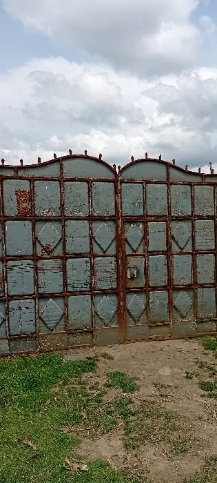 A two-panel, double-leaf metal gate, exhibiting significant rust and peeling light blue-grey paint, stands centered in the frame in Heni Village, Kenya. The gate features a grid construction composed of primarily rectangular and central diamond-shaped panels. Each half of the gate is topped by a distinct arched section with multiple small, pointed finials. The ground immediately in front of the gate consists of a mixed surface of sparse green grass and exposed brown dirt. Above the gate, the sky is partially overcast with a mix of white and grey cumulus clouds. No individuals, animals, or active events are present within the scene.