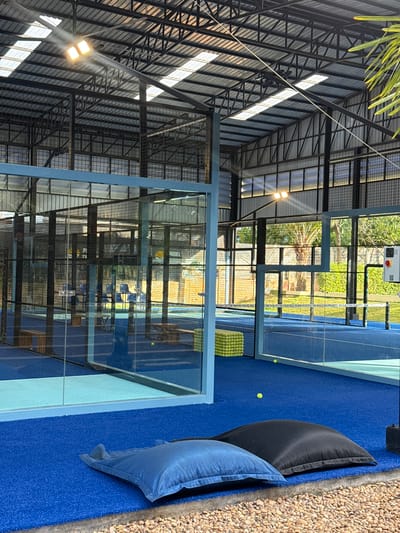 This media file depicts an outdoor padel tennis court in Ban Rak, Thailand. The scene features a modern padel court with blue turf and glass walls, enclosed by metal fencing. Outside the court, there are several large, dark-colored cushions, suggesting a seating or relaxation area. The court is situated in a well-lit environment, likely during the daytime, with sunlight filtering through the structure's metal roof and illuminating the playing surface.
The court appears to be set up for play, with a stack of bright yellow padel balls visible near one of the goals. A few individual balls are scattered on the court and the surrounding blue turf. In the background, lush green foliage and trees are visible, indicating a tropical or subtropical climate, consistent with the Thailand location. The architecture of the surrounding structure, with its exposed metal beams and corrugated roof, suggests a functional, perhaps sports-oriented facility.
There are no people visible in the media file, so no activity or event involving participants can be directly observed. However, the presence of the court and equipment strongly implies that padel tennis is the intended activity. The overall mood of the scene is tranquil and inviting, with the bright colors of the court and the natural greenery creating a pleasant atmosphere. The weather appears to be clear and sunny.