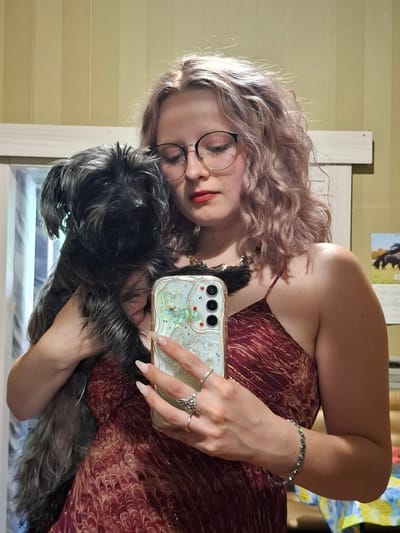 The image shows a young woman in Donetsk, Ukraine, taking a selfie with her dog. She is wearing a red and gold patterned dress and has blonde, curly hair. She is also wearing glasses, red lipstick, and several rings on her fingers. Her dog, a small black and gray breed, is held in her arms. The background appears to be an indoor setting with light yellow striped walls. There is a calendar visible on the wall with an image of a horse. The lighting suggests it is daytime. The woman seems happy and relaxed, interacting with her dog.