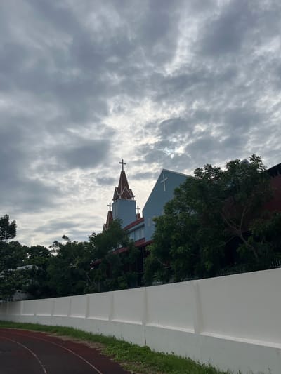 A low-angle shot captures a church structure with multiple steeples, each topped with a cross, against a cloudy sky. The building's lower half is painted a light blue with red trim, while the upper portions of the steeples are red with white accents. Trees with lush green foliage partially obscure the building, creating a layered effect. In the foreground, a white wall runs horizontally across the frame, and a portion of a red running track is visible to the left. The overall scene suggests a humid, overcast day in Phan Thiết, Vietnam. No people are visible in the image.