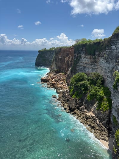 The image captures a coastal scene in Uluwatu, Indonesia, viewed from a high vantage point. A steep, rocky cliff dominates the landscape, with lush green vegetation clinging to its upper sections. The cliff face is textured with layers of rock and earth, descending into the turquoise waters of the ocean. The water is clear near the shore, revealing underwater rocks and the seabed, then deepens in color further out. The sky is a bright blue, scattered with puffy white clouds.