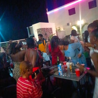 This image shows a crowded outdoor gathering at night in Ube, Nigeria. The scene appears to be a party or social event, with a large group of people socializing around a table. The table is covered with glasses, red plastic cups, and bottles, including what looks like a liquor bottle. Several people are standing or leaning around the table. There is a fair amount of ambient lighting. In the background, there is a modern building with a lit-up exterior, possibly indicating a bar or entertainment venue. The sky is dark, suggesting it is either late evening or night.