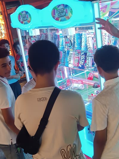 In Viadana, Italy, a group of young men are gathered around an illuminated claw machine, likely in an arcade or entertainment venue. The machine, featuring a bright turquoise top with "TOY CLAW" branding, is filled with various cylindrical plush toys. Several individuals are observing the game; one young man in the foreground, facing away from the camera, wears a white t-shirt with a black crossbody bag. To his left, another young man in a white polo shirt is looking towards the machine. In the background, two other young men are also watching the game, one with a surprised expression. The interior of the claw machine is visible, showcasing the prizes and the mechanics of the game. The lighting is predominantly from the machine itself, creating a vibrant and engaging atmosphere.