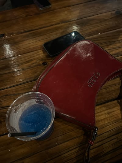 The scene captures a close-up, overhead view of items resting on a dark, reflective wooden table surface, indicative of a casual dining or bar setting in Wombarra, Australia. In the foreground, positioned to the lower left, is a clear plastic cup containing a bright blue, semi-frozen beverage with a black plastic straw inserted. Adjacent to the cup, occupying the right side of the frame, lies a shiny red purse or clutch with a metallic silver "GUESS" logo embossed on its front. Behind these items, two dark-colored smartphones are visible lying flat on the table, one partially obscured in the upper left, and another positioned directly behind the red purse. No individuals are depicted, and no specific actions or interactions are in progress. The wooden table exhibits visible grain and reflections, suggesting a polished or possibly damp surface.