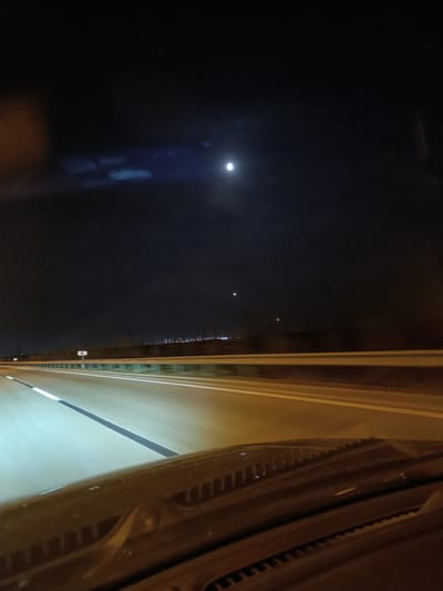 The image captures a nighttime scene from the perspective of a vehicle traveling on a highway. The road ahead is illuminated by headlights and streetlights, with white lane markings visible. A guardrail runs along the side of the highway, separating it from the dark landscape beyond. In the distance, a faint line of city lights suggests the presence of a populated area. The sky is dark, with a bright moon prominently visible. There are also two other faint lights in the sky, possibly stars or distant aircraft. The foreground of the image shows the dashboard of the vehicle, with its textured surface and a hint of the windshield. The overall impression is one of a solitary journey on a highway at night, under a moonlit sky. The time of day is clearly night. The weather appears clear, allowing for good visibility of the sky and distant lights. The location is identified as Surakhany Raion, Azerbaijan, suggesting a highway setting in that region. No people are visible in the image.