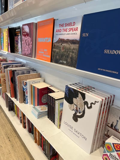 The image displays an interior view of a bookstore or library in Brisbane City, Australia, with multiple shelves filled with books. The perspective is from slightly above, looking down across several white shelves packed with publications. Books are arranged in horizontal stacks, showcasing their covers, and also standing vertically, displaying their spines. The scene suggests a retail environment for browsing or purchasing books.

A significant portion of the visible collection focuses on Indigenous Australian art, culture, and history. Prominent titles include "SHADOW ART OF THE SPINIFEX PEOPLE," "SUN," and "THE SHIELD AND THE SPEAR THE KIMBERLEY LAND COUNCIL" by Joe Fox. Other books with Indigenous themes include "IWANTJA IRITI KUWARI TITUTIARA," "KUNTJI TEXTILES," "BALGO Indigenous Art," and "JUNIOR ATLAS INDIGENOUS." A distinctive white book, "LOUISE SAXTON cut & pinned," featuring an intricate eagle and snake design, likely pertains to art.

Other categories of books are also present, such as several copies of "HEALTH" and "INNOVATION." General titles like "YOU CALL IT" and "Once" are visible. On lower shelves, books by Indigenous author "ANITA HEISS" and "HEART" can be seen. Partially visible titles further reinforcing the Indigenous focus include "First Nation," "BRENDAN PASCOE" (likely referring to "Dark Emu"), and "WELCOME TO THE LAND." The bright, even lighting suggests an indoor daytime setting. The collection reflects a diverse range of subjects, with a notable emphasis on Australian and Indigenous cultural narratives and arts.