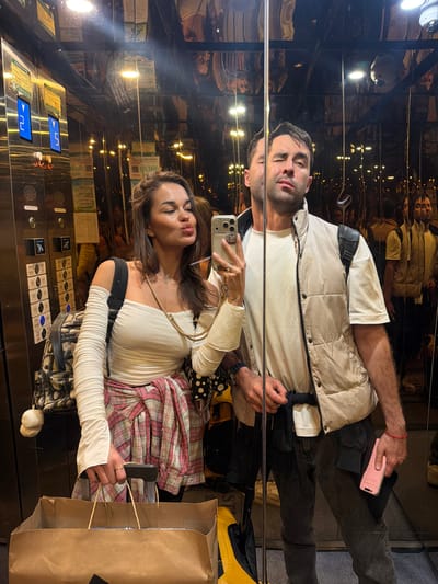 Two individuals are visible in an elevator with mirrored walls, capturing a selfie. The woman holds a smartphone in her right hand, her lips pursed for the photo. She wears a light-colored top, long sleeves, and a pink plaid skirt. A shopping bag is visible to her right. The man stands to her left, also visible in the mirrored reflection. He wears a light-colored shirt, a vest, and dark pants. His right hand holds a pink phone. The elevator's control panel is visible, and the reflection shows the city environment, possibly Zhangjiajie, China, due to the unusual elevator location and appearance. The light inside the elevator is dim, but the reflection reveals a brightly lit street scene.