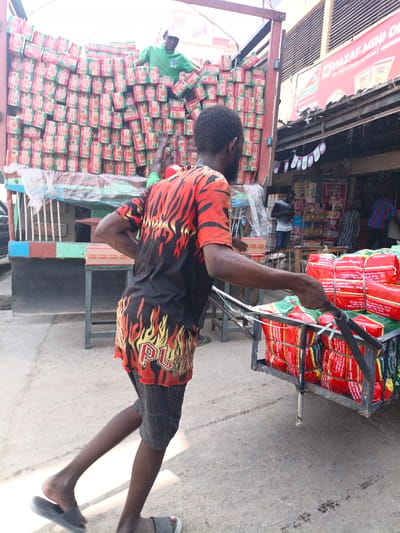 The image captures an active street-level commercial scene in Fagge C, Nigeria. In the foreground, a dark-skinned male, facing away from the viewer, is propelling a metal handcart. He wears a black t-shirt with red and yellow flame graphics, plaid shorts, and grey slide sandals. The handcart is loaded with multiple bundles wrapped in red material, secured with white strapping; text on the bundles partially reads "NEW HOP INTERNATIONAL TRADE".
In the midground, a large truck is parked, its bed filled with numerous stacked cardboard boxes featuring a distinct green and pink/red pattern. A dark-skinned male, wearing a green t-shirt and white cap, stands atop these boxes within the truck, actively handling them. Another individual's leg, also in green attire, is partially visible near the truck's rear.
The background features a storefront with a prominent red sign partially legible as "MAZAF MINI D...". The store's interior displays various packaged goods. Several individuals are discernible within or near the store's entrance. The ground is paved, showing tire marks and general wear. Shadows suggest outdoor daylight conditions.