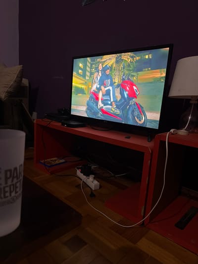 An indoor scene in Vila Real, Portugal, depicts a dimly lit residential room, illuminated primarily by a flat-screen LG television displaying a vibrant image. The television screen shows a man and a woman seated on a red scooter. The man, in a dark hoodie with white stripes and yellow-soled shoes, is at the front, with the woman, in a light-colored top, behind him. The background suggests an urban night setting with illuminated buildings and palm trees.
The television rests on a red, open-shelf entertainment unit. A black electronic device, possibly a media player, is situated on this unit below the screen. To the right, an identical red open-shelf unit supports a table lamp with a white fabric shade. To the far left, a portion of a light-colored cushion from a sofa or armchair is visible, adjacent to a vertical appliance. Numerous cables are visible, mostly tangled below and behind the red entertainment unit, connecting to a white power strip with several European-style plugs on the parquet wood floor. In the immediate foreground, a white mug with dark, partially visible lettering ("E PAR REPET") rests on a dark surface, likely a coffee table. The walls are painted a dark, possibly purple, color.