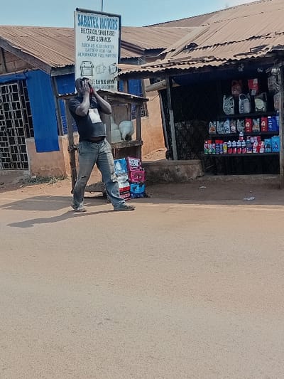 The image depicts a street scene in Ayangba, Nigeria, under clear daylight conditions. A single male subject, dark-complexioned, stands centrally on an unpaved road surface. He is wearing a dark t-shirt and light-colored trousers. His hands are raised, holding a red mobile phone to his right ear, thereby obscuring his face. A distinct shadow is cast to his left.
Behind the subject and to his left, a single-story building with a corrugated metal roof and blue window frames is visible. A prominent vertical sign affixed to a pole in front of this building reads "SABATEX MOTORS" at the top, followed by "ELECTRICAL SPARE PARTS SALES & SERVICES," with a list of items including "KICK STARTER," "BATTERY," and "FUEL PUMP."
To the subject's right, an adjacent single-story structure also with a corrugated metal roof functions as a retail establishment. Its open storefront displays multiple shelves stocked with various bottles of motor oil and other automotive fluid containers; "JTT" branding is visible on some products. Several stacked boxes, some appearing red or pink, are on the ground near the shop entrance, to the right of the subject. A white, indistinct object is positioned behind the subject, near the entrance of the right-hand shop.