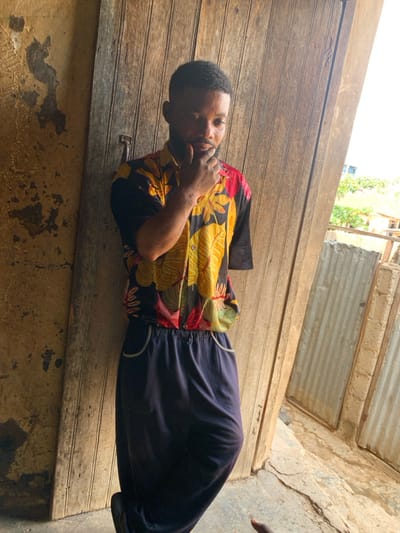 A young male, with a short beard and dark hair, stands in what appears to be an open doorway or alcove in Okene, Nigeria. He wears a short-sleeved button-up shirt with a black base and vibrant yellow, red, and green floral patterns, paired with dark blue wide-leg trousers featuring white piping around the pockets. His right hand is raised, with fingers touching his chin/mouth area. His left arm is not fully visible, appearing to terminate above the elbow. To his immediate left is a weathered wooden plank wall or door, secured with a visible metal handle. Further left, an adjacent wall shows significant peeling of light-colored plaster. To his right, beyond the doorway, vertical corrugated metal sheets are visible, partially obstructed by concrete blocks. Distant greenery and a light-colored building are discernible in the background. The ground beneath him is an uneven concrete or packed earth surface.