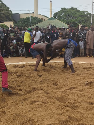The image captures a traditional wrestling match in an outdoor arena in Kafanchan A, Nigeria. The wrestling ring is a large sandy area enclosed by a white boundary line. Two shirtless men are engaged in wrestling, one wearing gray shorts and the other wearing blue and red shorts. They are surrounded by a large crowd of spectators, including men, women, and children. Many in the crowd are standing, while some are seated on the ground. There is a building with a green roof and a white minaret in the background. Trees and overcast skies are visible above the crowd.