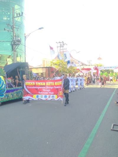 An outdoor parade scene unfolds on a paved street in Madiun, Indonesia. In the foreground, a dark gray asphalt road occupies the lower third of the frame, featuring a distinct green line extending longitudinally on the right side. A red and white banner, held horizontally across the road by two individuals, is central. The banner displays yellow text "MKKS SMKN KOTA MADIUN" and white text "Keanekaragaman Budaya Nusantara Menuju Indonesia Maju" (Diversity of Nusantara Culture Towards a Developed Indonesia), along with a red circle containing the number "16" and four small icons.

The individual holding the banner on the right wears a dark jacket with gold accents, dark pants, and a dark cap. To the left of the banner, a decorative float, primarily green and blue with peacock-eye patterns, moves along, carrying at least two partially visible individuals.

Behind the banner, a uniformed group marches in formation. This group, consisting of approximately 10-15 individuals, wears white long-sleeved uniforms with red detailing and white headwear. Several members carry poles with flags, including the red and white flag of Indonesia and multiple blue and white flags with a distinct crest. Following this group, a longer procession of individuals dressed in various traditional Indonesian attire proceeds down the street.

In the background on the left, a tall building with a curved, reflective green glass facade is prominent. A utility pole with multiple cross arms, transformers, and a network of overhead wires stands in front of it. A decorative street lamp extends over the road from the left. On the right background, buildings with orange-tiled roofs and green trees are visible, leading to a festive archway spanning the road. This archway is decorated with red and white fabric and green elements, displaying the text "MADIUN Carnival." The sky is intensely bright and overexposed, indicating strong overhead sunlight.