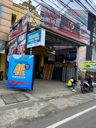 The image shows a street scene in front of a printing shop in Kecamatan Margahayu, Indonesia (latitude: -6.976, longitude: 107.566). The shop is called "Cahya Gradika" and offers printing services like print UV, DTF, and spanduk (banners).  A blue sign in front of the shop advertises a 40% discount on printing services, valid until 10 June 2023. Two men are sitting on the sidewalk in front of the shop, one wearing a yellow shirt and the other wearing a black shirt, both looking at a mobile phone. A motorcycle is parked behind them. The street is paved with concrete, and there are power lines and wires running across the top of the image.  The building is yellow with a brown roof and is adorned with various other signs.
