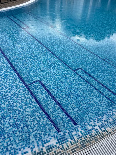The image shows a close-up view of a swimming pool with blue mosaic tiles. Several steps are visible, leading into the water. The water's surface is disturbed by ripples, suggesting it is raining. The poolside is tiled and there is a metal grate drain visible. The location is the swimming pool at the hotel "Interhotel Sandanski" in Sandanski, Bulgaria.