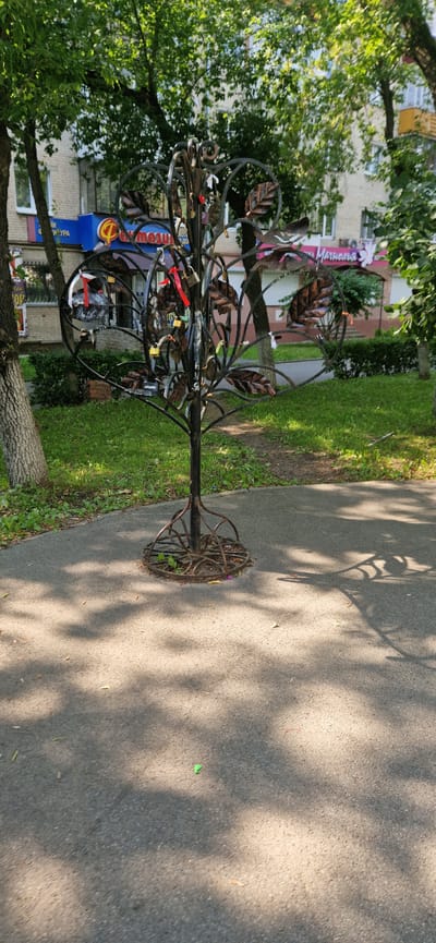 A metal sculpture resembling a tree stands in a park area with a paved walkway. The tree sculpture is adorned with numerous padlocks and ribbons, likely representing love locks. Behind the sculpture, a grassy area is visible, bordering a building with commercial storefronts. Trees with green foliage are present in the background, casting dappled shadows on the walkway. The scene appears to be outdoors, under natural daylight. The location is identified as Miass, Russia.