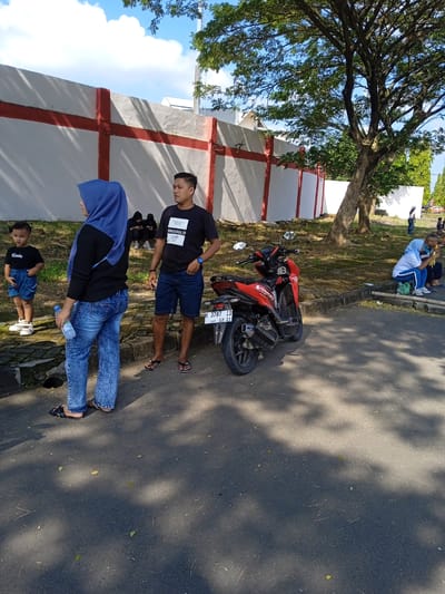 The image depicts an outdoor scene in Kendal, Indonesia. Several people are present, including a woman in a blue hijab and jeans, a man in a black T-shirt and shorts, and a young boy in a black T-shirt and blue shorts. The woman is holding a water bottle. A red motorcycle is parked nearby. A white wall with red accents is in the background. A large tree is also visible. Other individuals are seated further away. The scene appears to be near a road with shadows cast on the pavement.