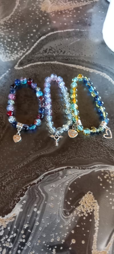 Three beaded bracelets with charms are laid out on a dark, marbled surface. The bracelets are of varying colors: one is predominantly blue with accents of red, purple, and pink; another is a light, translucent purple; and the third is a mix of yellow, green, and blue. Each bracelet has a silver-toned charm attached. The leftmost bracelet has a heart-shaped charm. The middle bracelet has a key-shaped charm with small wings. The rightmost bracelet has a heart-shaped charm with the inscription "Thank You." The surface appears to be a tabletop with a swirling, textured pattern. There is no visible indication of the city of Iskra, Bulgaria, or any specific location. The image is a close-up shot, focused on the bracelets and the surface they are on. No people or actions are depicted.