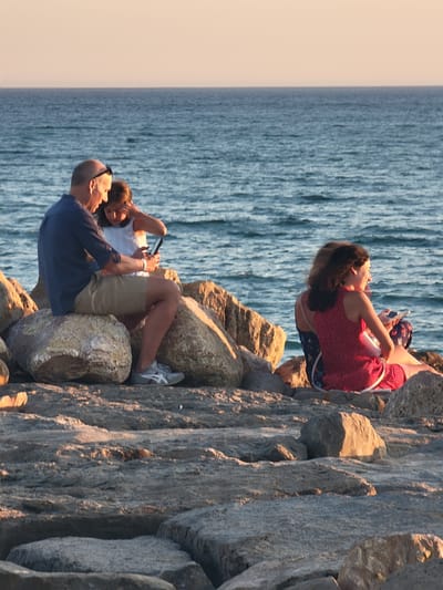 In Quarteira, Portugal, two groups of people are seated on rocks by the sea. In the foreground, a man in a dark blue shirt and tan shorts sits on large rocks, looking down at his phone, with a young girl in a white top beside him, also looking at a phone. To the right, a woman in a red dress and another person in a patterned dress sit on rocks, with the woman in red also looking at a phone. The sea is visible behind them, with a clear sky above. The rocks appear to be part of a coastal fortification or breakwater.