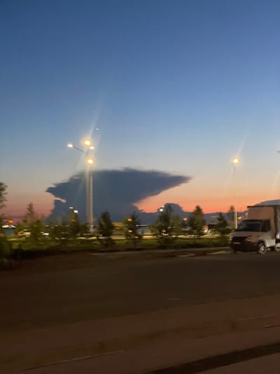 The image depicts a twilight scene in Kokshetau, Kazakhstan. A distinctive cloud formation, resembling a human profile with a prominent nose and chin, dominates the sky. In the foreground, a road is visible with a moving van and a streetlight with multiple illuminated bulbs. A line of trees and streetlights runs along the roadside, and in the distance, lights from buildings or a cityscape can be seen. The sky transitions from a deep blue at the top to shades of orange and pink near the horizon, indicating the setting or rising sun. The overall impression is a serene urban landscape at dusk, punctuated by the unusual cloud formation.